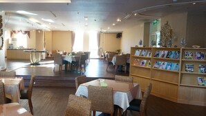 Ein Hotel-Essbereich mit Tischen und Stühlen, im Hintergrund eine Küche und ein Bücherregal mit Zeitschriften.