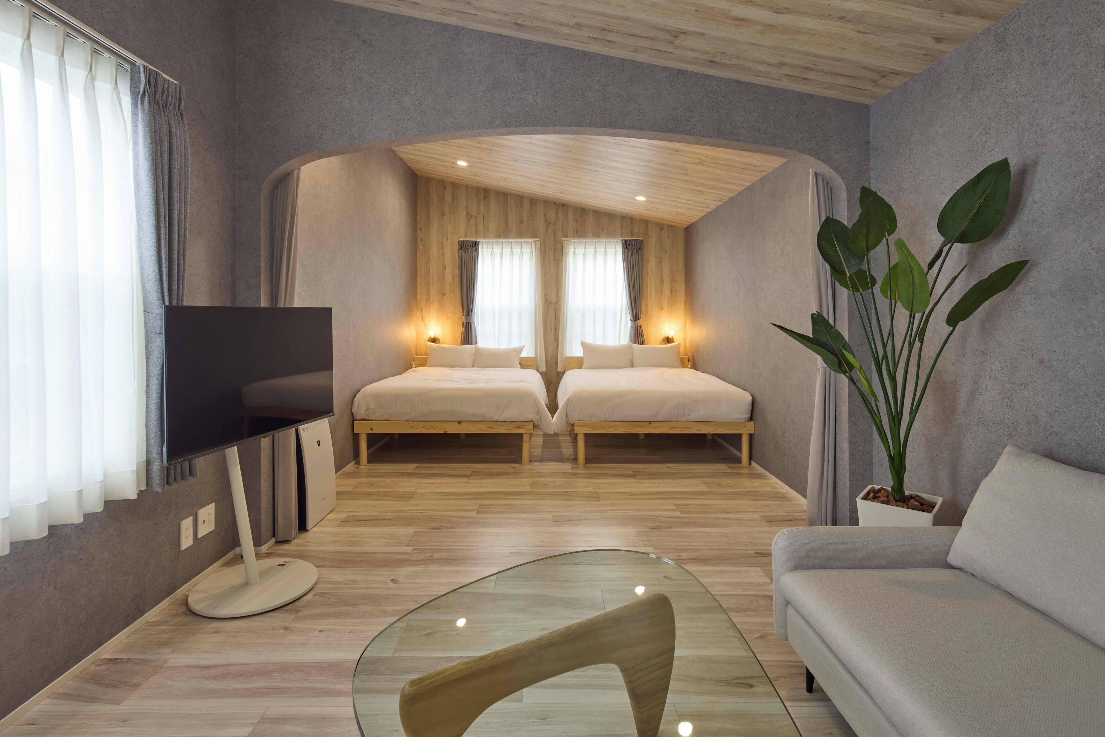 A modern bedroom with two beds, a TV, a glass table, and a potted plant.