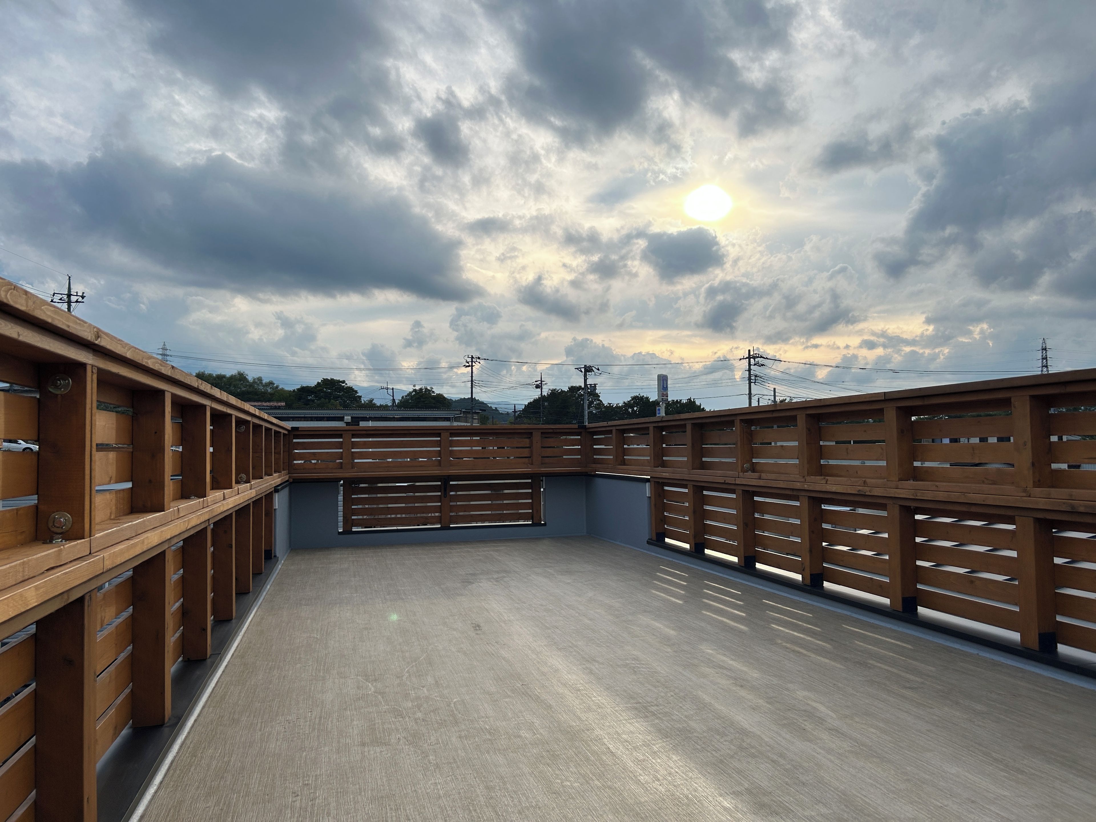 A covered outdoor area with a wooden fence and a concrete floor, under a cloudy sky with the sun visible.