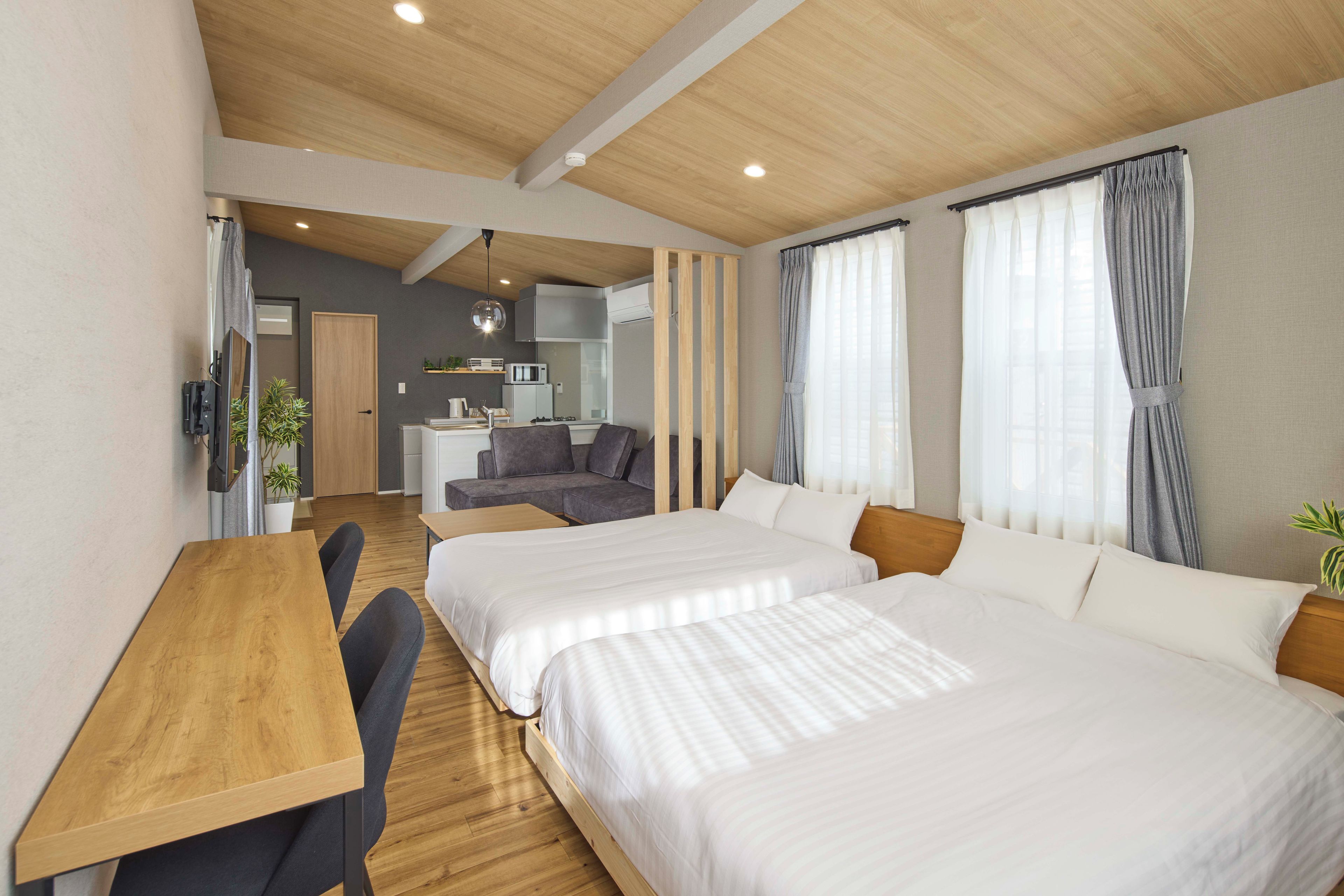 Habitación de hotel con dos camas, un escritorio de madera, una kitchenette y un sofá gris.