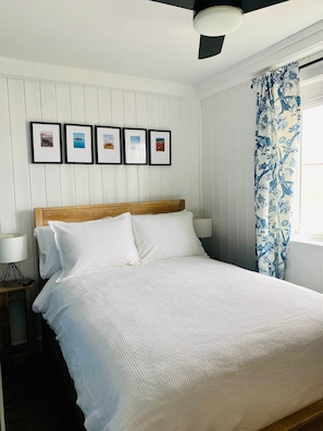 A bedroom with a bed, white walls, a ceiling fan, and framed pictures on the wall.