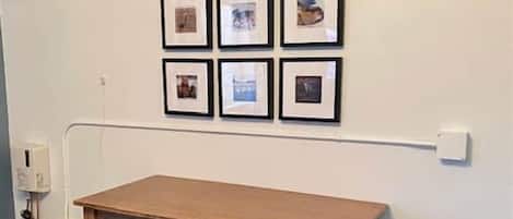 A room with a wooden table and two blue stools, a wall-mounted power outlet, and a white wall with framed pictures.