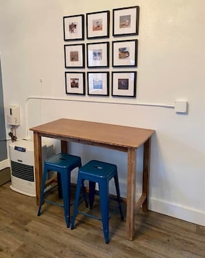 Ein Zimmer mit einem Holztisch, zwei blauen Hocker, einer Wandsteckdose und einer weißen Wand mit gerahmten Bildern.