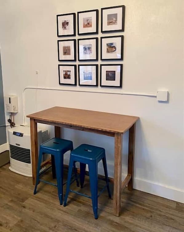 A room with a wooden table and two blue stools, a wall-mounted power outlet, and a white wall with framed pictures.