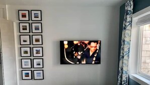 Huoneessa on seinälle asennettu televisio, pieni pöytä ja seinällä useita kehystettyjä tauluja.