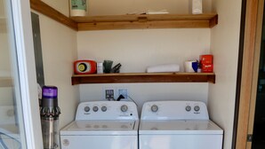 A compact laundry room with two Whirlpool dryers, a shelf with various items, and a broom.