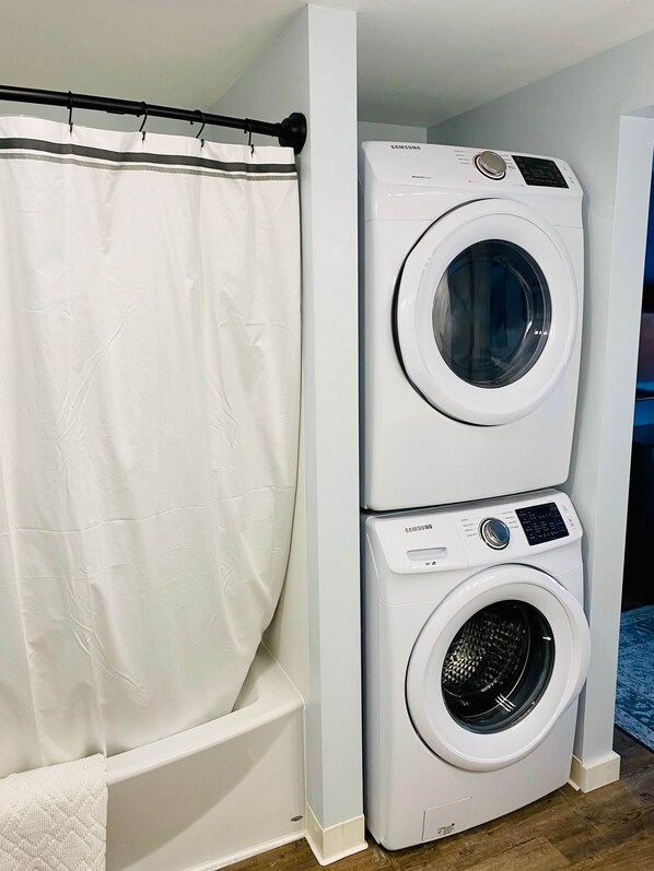 A stacked washer and dryer unit in a room with a white shower curtain and a wooden floor.