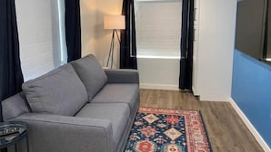 Ein Zimmer mit grauem Sofa, einem gemusterten Teppich, einer Stehlampe und einem an der Wand befestigten Flachbildfernseher.