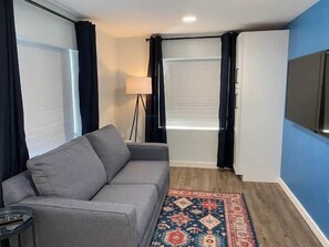 A room with a grey sofa, a patterned rug, a standing lamp, and a flat-screen TV mounted on the wall.