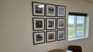 Een kamer met een ronde tafel en twee bruine stoelen, een raam met uitzicht op groen en een muur met ingelijste foto's.