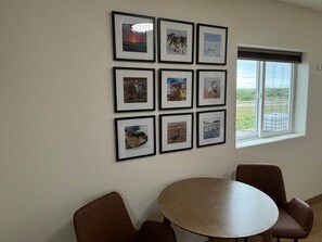 Ein Zimmer mit einem runden Tisch und zwei braunen Stühlen, ein Fenster mit Blick auf Grünflächen und eine Wand mit gerahmten Bildern.