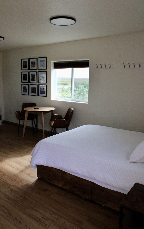 A room with a large bed, a dining table with chairs, a window with a view, and a wall with framed pictures.