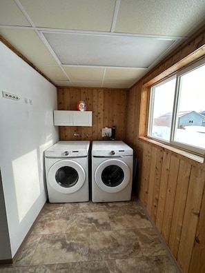A laundry room with two white washing machines, a cabinet above, and a window to the right.