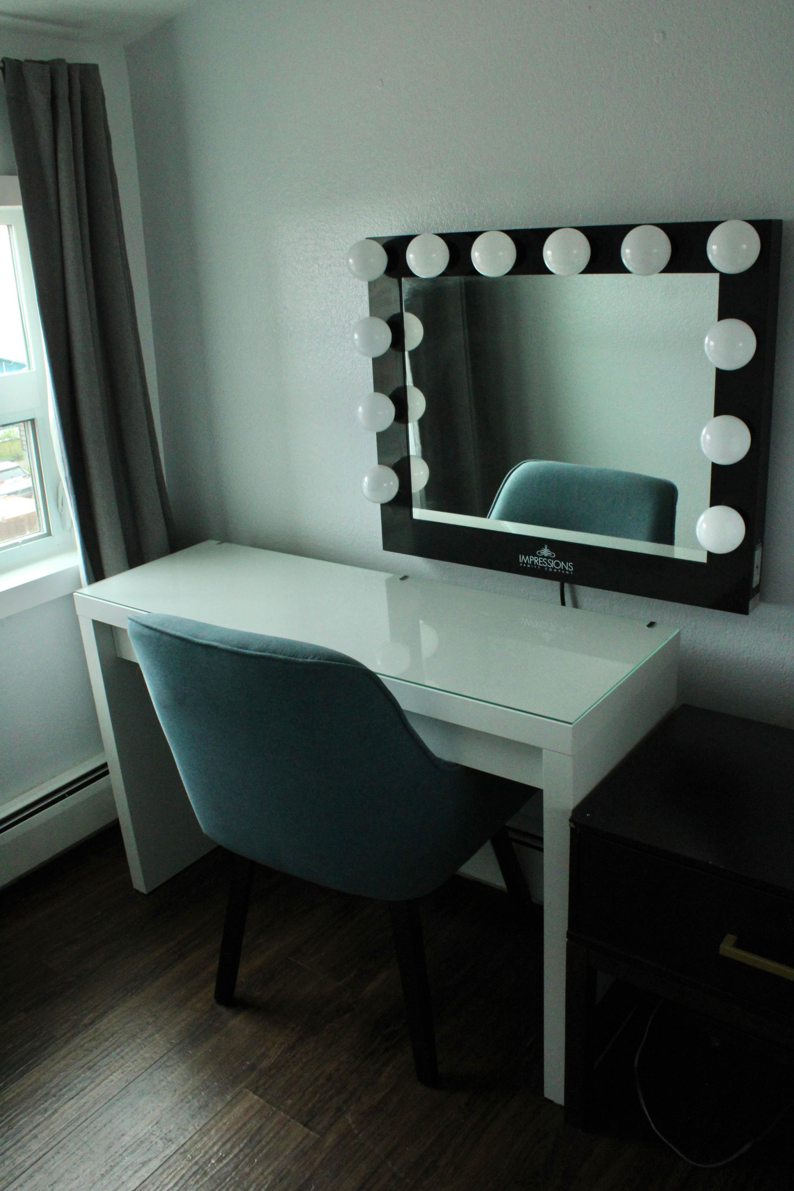 Een kamer met een wit bureau, een blauwe stoel en een spiegel met verlichting aan de muur.