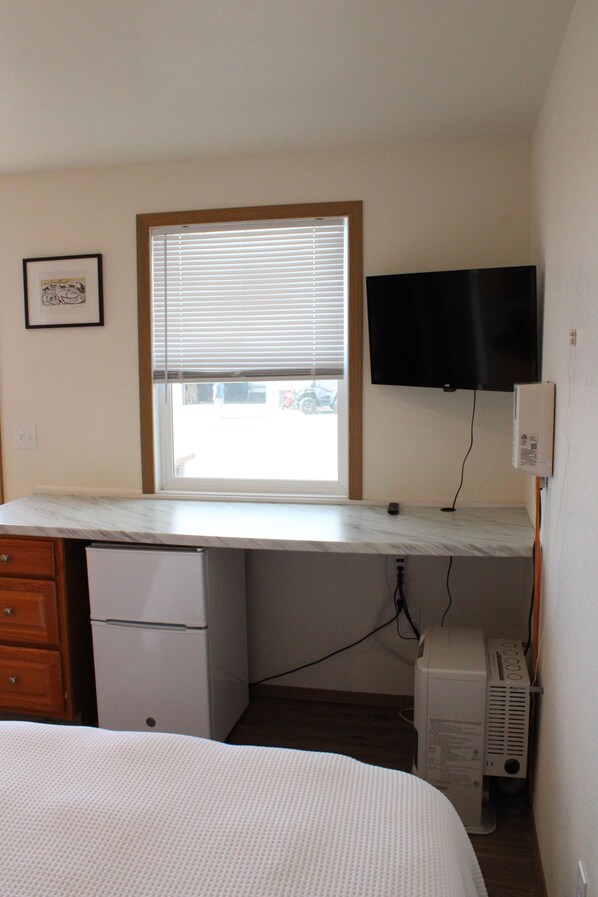 A bedroom with a bed, a desk with a computer, a television, and a window with blinds.