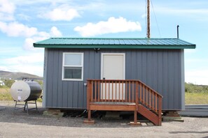 A small, single-story building with a green roof, a wooden staircase, and a metal tank outside.