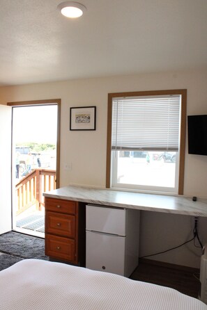 A bedroom with a bed, a desk, a dresser, a window with blinds, a framed picture on the wall, and a ceiling light.