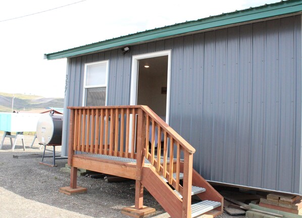 A small building with a wooden staircase leading to a door, a barbecue grill, and a table with a blue cover outside.