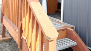 Um deck de madeira leva a um edifício com fachada de metal e uma porta aberta para um quarto com cama.