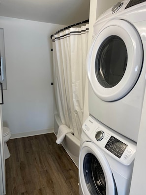 A laundry room with stacked washers and dryers, a shower curtain, and a wooden floor.