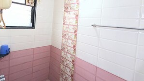 Ein Badezimmer mit weißen und rosafarbenen Fliesen, einer Dusche mit floralem Trennelement, einem Fenster mit Ventilator und einem blauen Eimer auf dem Boden.