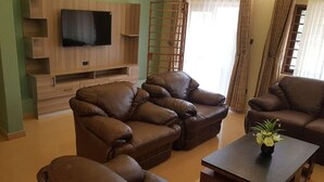 A living room with a flat-screen TV mounted on a wooden wall unit, leather sofas, a coffee table with a plant, and a window with curtains. - Cactus Hill Side Hotel (Gulu)