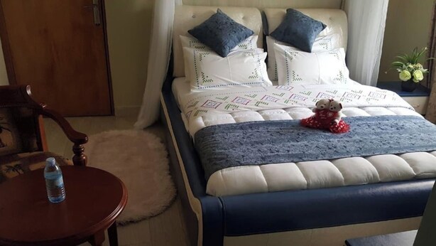 A bedroom with a bed, pillows, a teddy bear, a water bottle, a chair, and a small table. - Cactus Hill Side Hotel (Gulu)