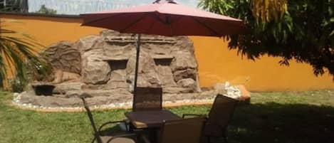 Un patio al aire libre con una mesa, sillas, un paraguas rojo y una pared de roca con un pequeño estanque.
