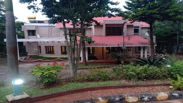 A two-story building with a red-tiled roof, surrounded by greenery and a paved courtyard.