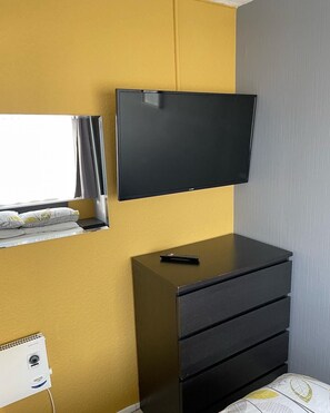 Habitación con una televisión de pantalla plana en la pared, un aparador negro con cajones, una ventana con cortinas y un pequeño escritorio blanco con una computadora.