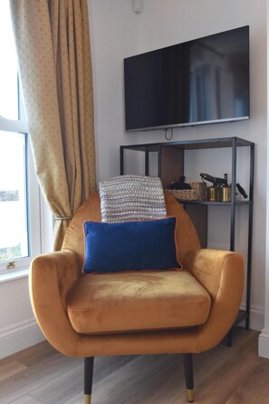 Una habitación con un sillón amarillo, una almohada azul, un televisor en la pared y una estantería con diversos objetos.