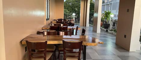 Un comedor con una larga mesa de madera y sillas, piso de baldosas y vista a áreas verdes.