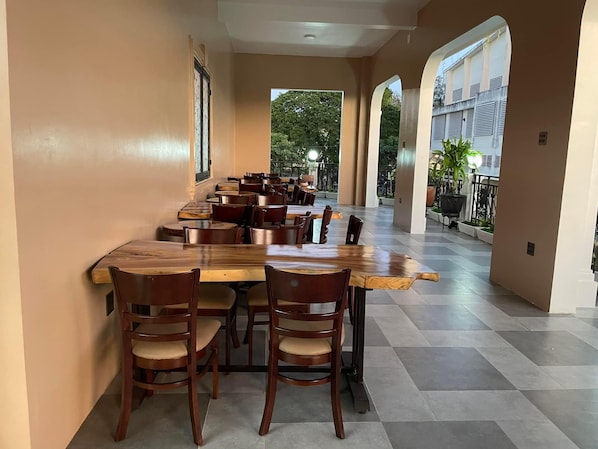 Uma área de refeições com uma mesa de madeira comprida e cadeiras, piso de azulejos e vista para o verde lá fora.
