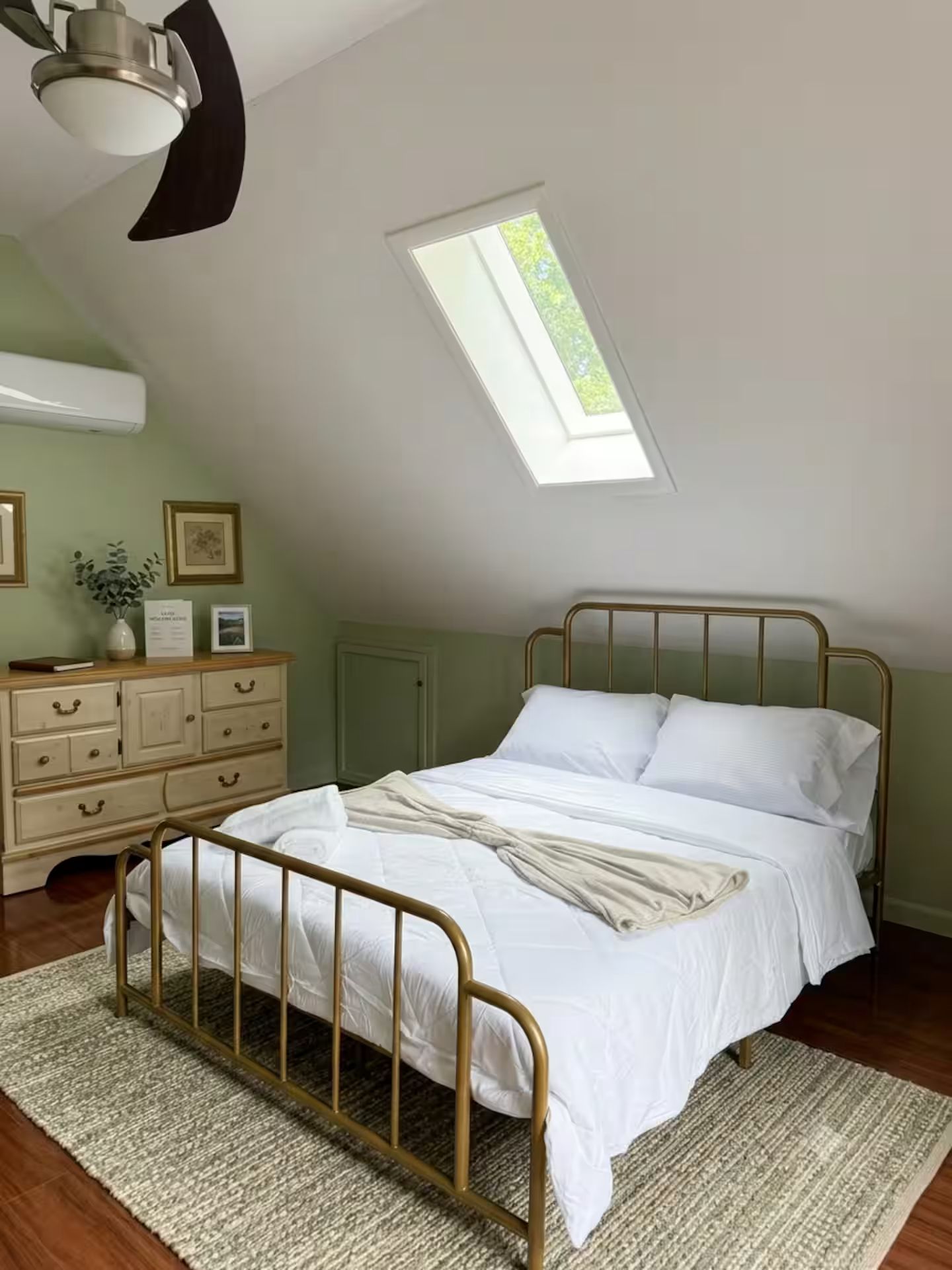 A bedroom with a sloped ceiling, a skylight, a bed with a metal frame, a dresser with drawers, and a small plant on the dresser.