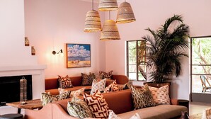 A cozy living room with a sofa, patterned cushions, a fireplace, and a large window with a view of greenery. - Sedia Riverside Hotel (Maun)