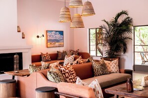 A cozy living room with a sofa, patterned cushions, a fireplace, and a large window with a view of greenery. - Sedia Riverside Hotel (Maun)