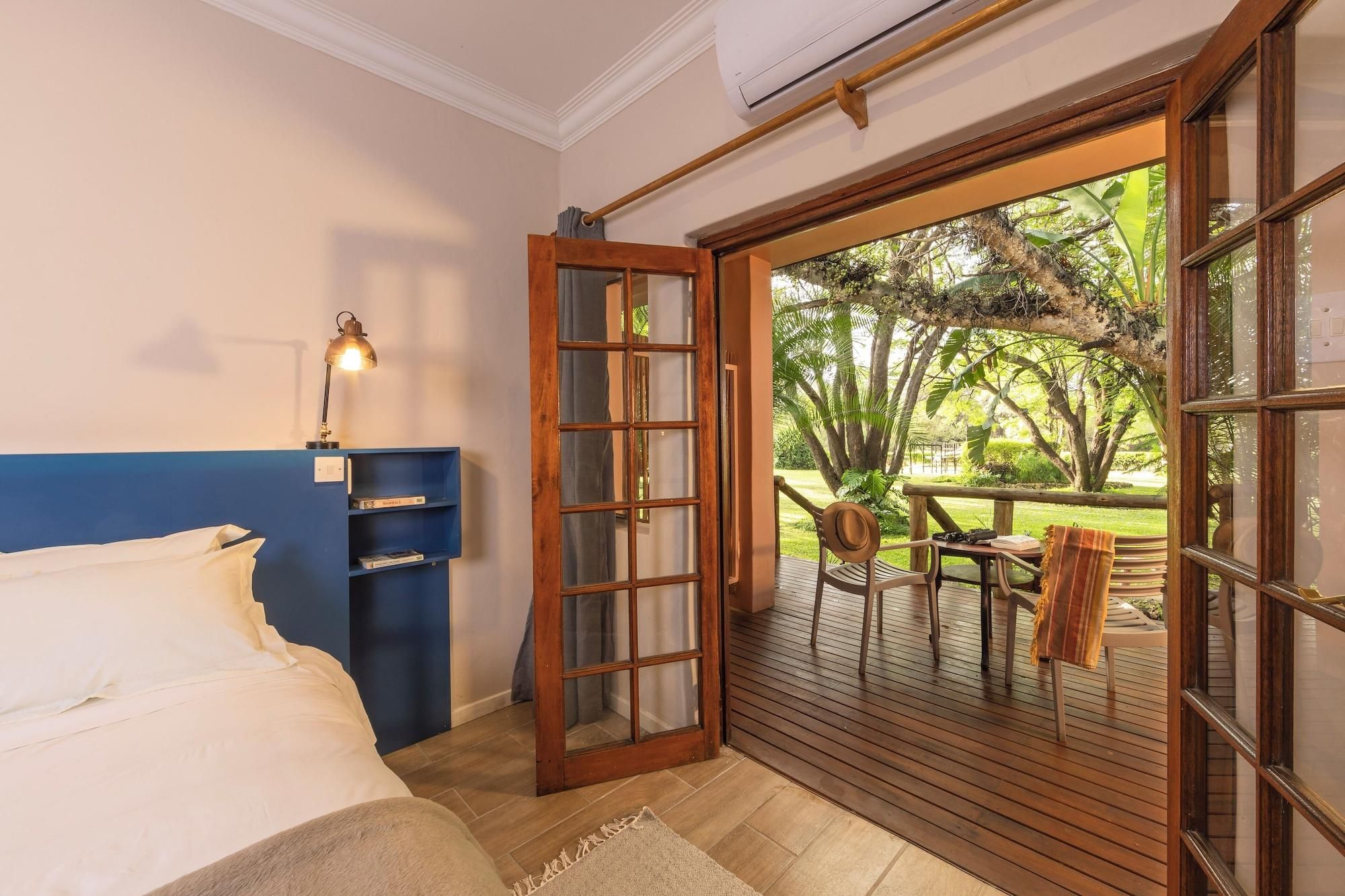 Un dormitorio con cama, un escritorio con lámpara, una estantería y un ventanal corredero que da a un área exterior con mesa y sillas.