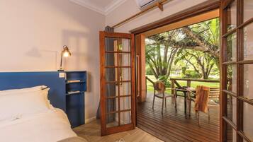 Un dormitorio con cama, un escritorio con lámpara, una estantería y un ventanal corredero que da a un área exterior con mesa y sillas.