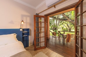 Un dormitorio con cama, un escritorio con lámpara, una estantería y un ventanal corredero que da a un área exterior con mesa y sillas.