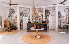 A lobby with a round table, wicker chairs, a reception desk, and a ceiling fan. - Sedia Riverside Hotel (Maun)