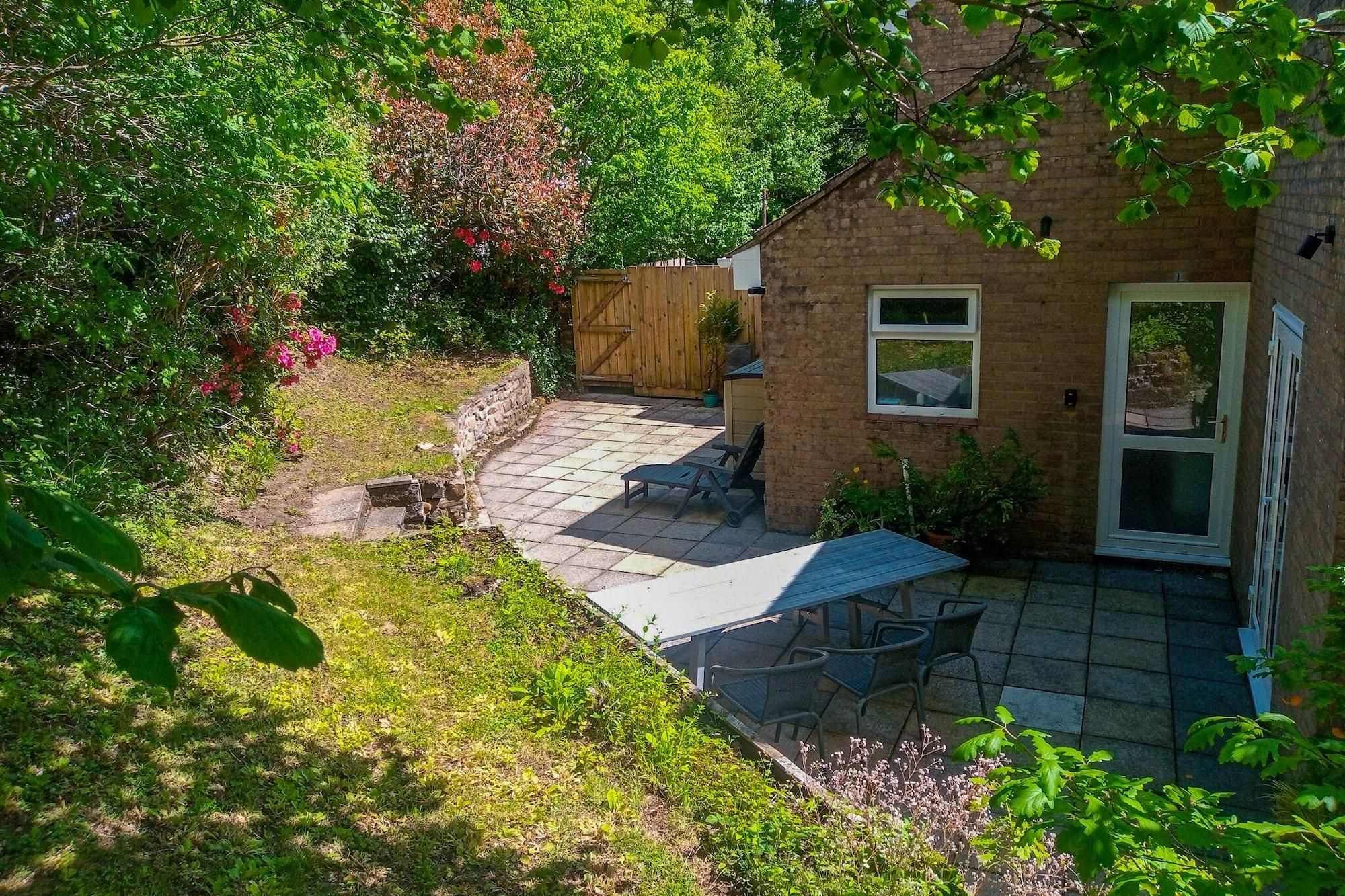 A brick house with a patio area featuring a table and chairs, surrounded by lush greenery and a wooden fence.