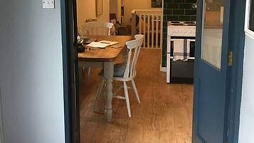 A dining area with a wooden table and chairs, a kitchen in the background, and a blue door.