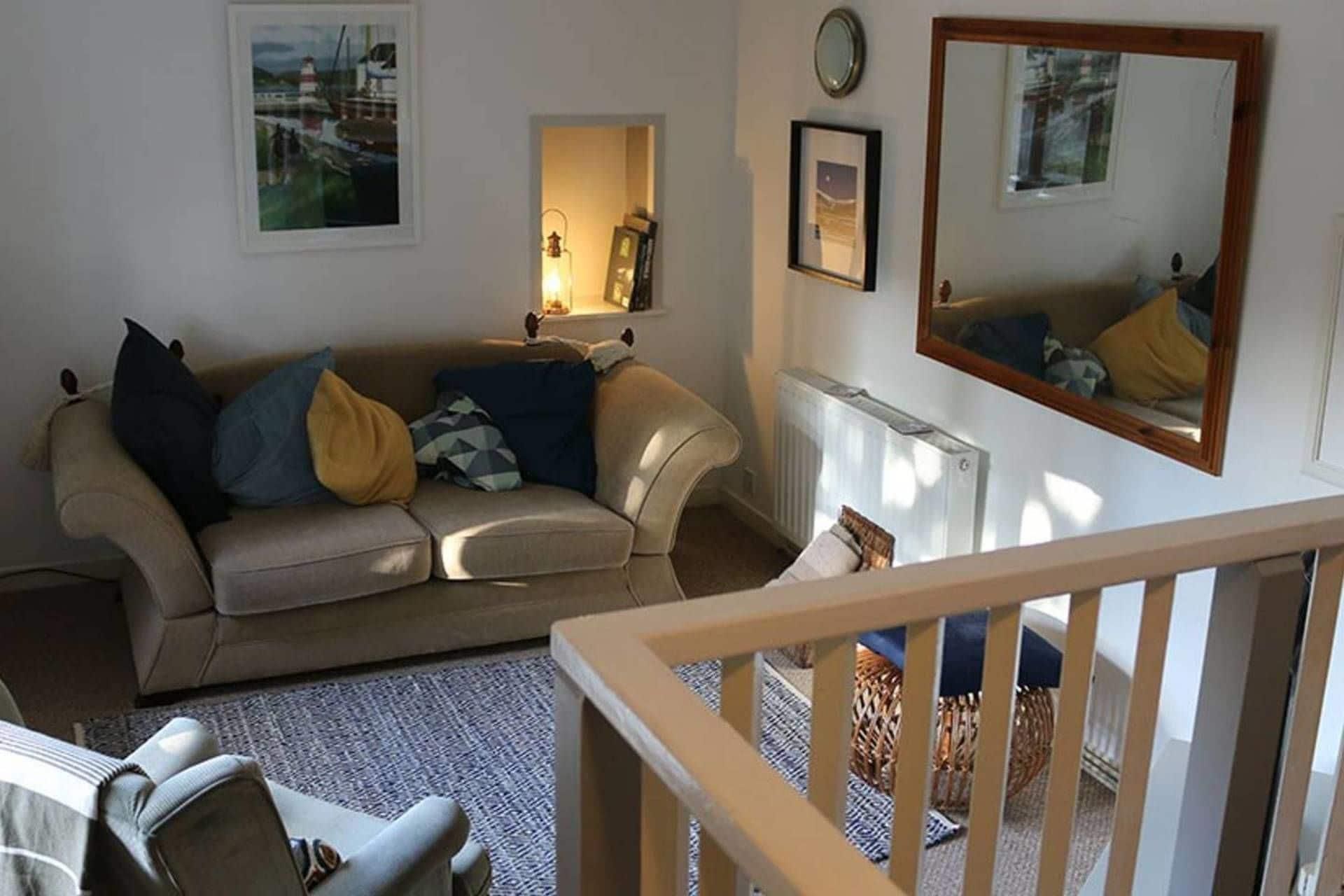 Ein Wohnzimmer mit einer Couch, einem Couchtisch und einem Sessel. Es gibt ein Fenster mit Blick auf Boote, eine Lampe und ein gerahmtes Bild an der Wand.