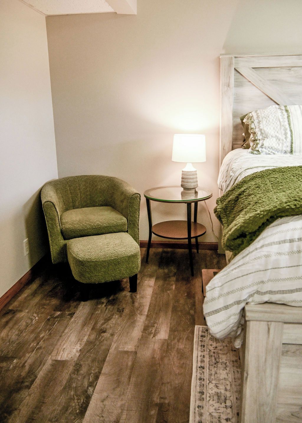 A room with a green armchair, a small round table with a lamp, and a bed with a green blanket.