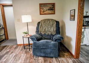 Ein Wohnzimmer mit einem blauen Sessel, einem kleinen Tisch mit einer Lampe und einem Bild an der Wand.