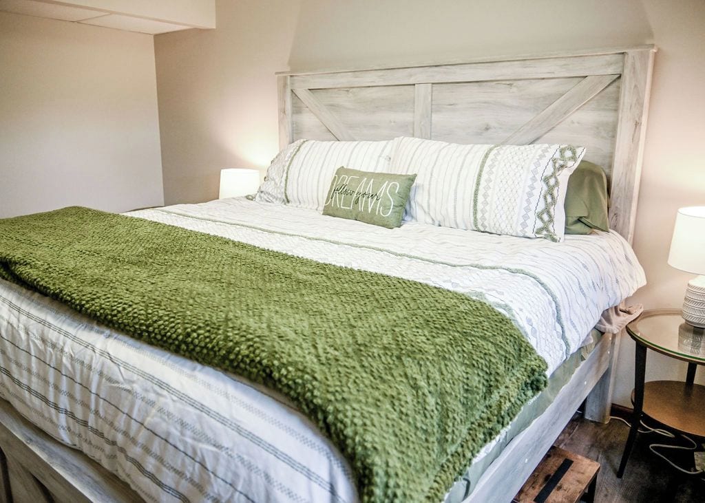 A neatly made bed with a green throw blanket, a pillow with the text 'Dreams,' and a wooden headboard.