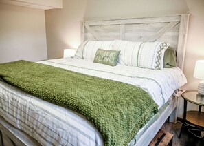 A neatly made bed with a green throw blanket, a pillow with the text 'Dreams,' and a wooden headboard.