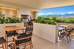 An outdoor dining area with wooden tables and chairs, a grill, and a view of a cityscape and mountains. - Chic Versalles Condo • Infinity Pool, Coworking (Puerto Vallarta)