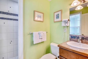 Une salle de bain comprenant des toilettes, un lavabo et un espace douche. Deux tableaux encadrés sont accrochés au mur, une serviette est suspendue à un porte-serviettes et un sèche-cheveux est fixé au mur.