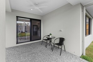 A patio area with a ceiling fan, a glass door leading to a kitchen, and a small round table with two chairs. - The Helensvale Haven | Modern 4BR Family Retreat (Helensvale)